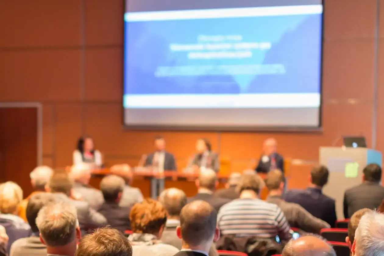 Audience attending a corporate panel discussion or conference session, with four blurred speakers seated on stage and a large presentation screen in the background.