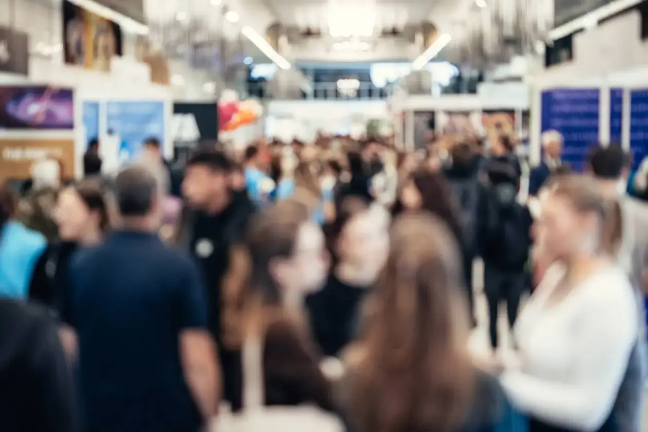 Blurred view of a busy indoor corporate event or trade show, with large crowds of people walking and talking in a well-lit exhibition hall.