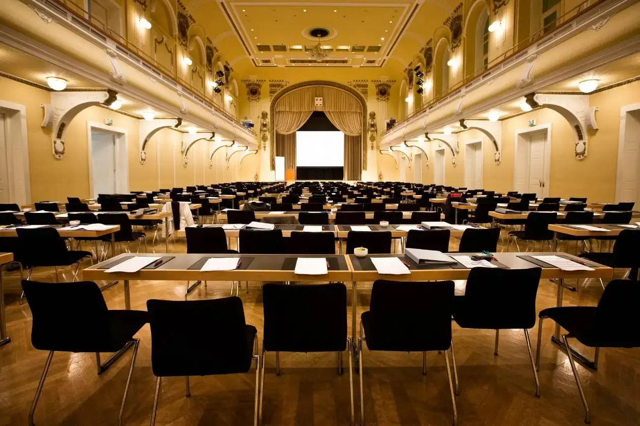 Large conference hall with ornate high ceilings, rows of black chairs and writing desks set up for attendees, and a projection screen on stage at the front.