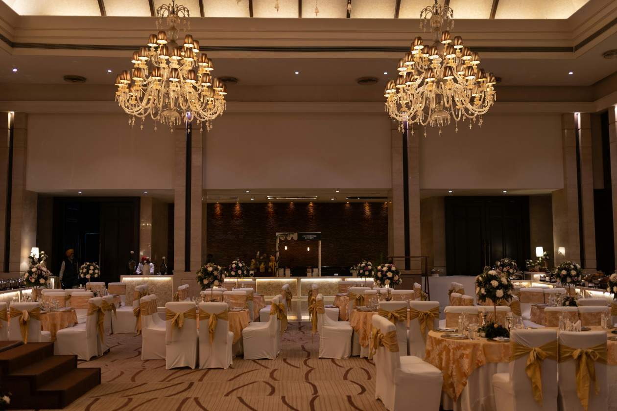 Elegant hotel ballroom prepared for an event, featuring round tables draped in gold and white linens, floral centrepieces, and chandeliers casting warm light across the room. The space includes a bar at the back and wide open floor areas designed for gatherings or presentations.