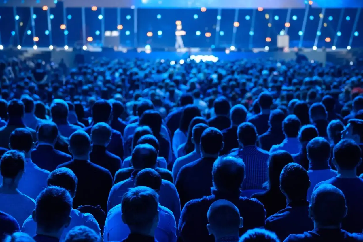 Large audience seated in a darkened conference hall watching a presenter on a brightly lit stage, with blue stage lighting creating an immersive atmosphere.