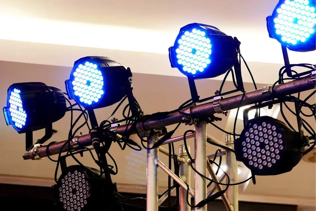 LED stage lighting fixtures attached to a metal truss during an indoor event setup, showing cables, mounts, and overhead positioning.